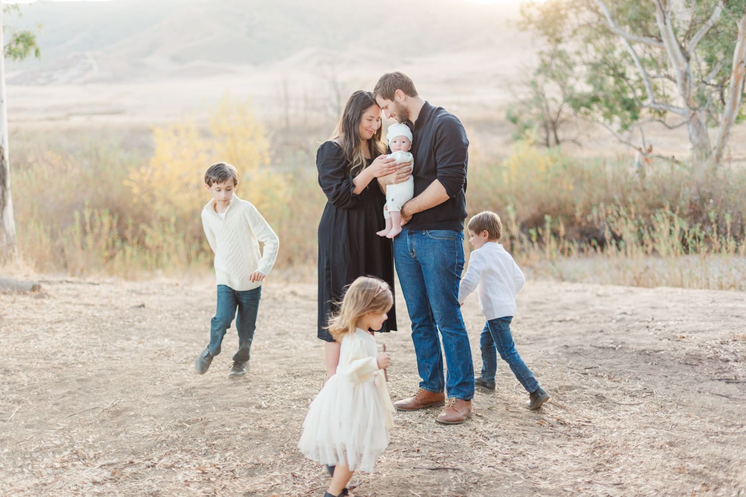 family photo, parents standing in the middle holing their baby while their three kids run around them