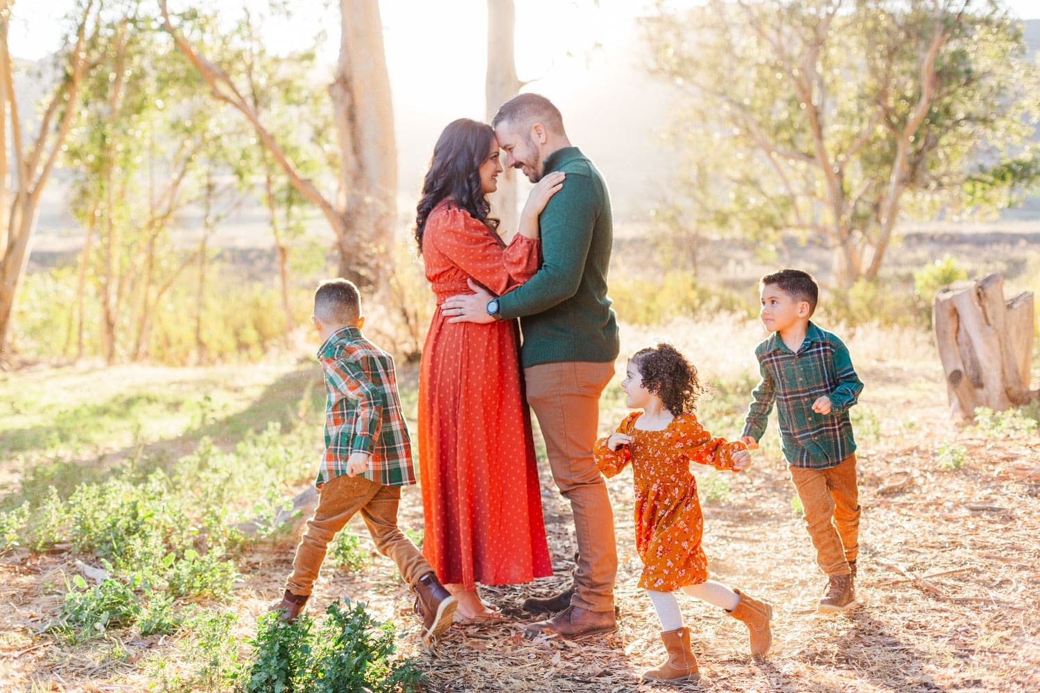 family photo, mom and dad hugging with children running around them, mom and daughter wearing an orange dress and dad and boys wearing green.