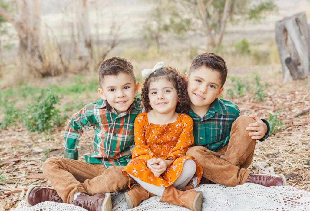 three children sitting on a blanket. boys wearing plaid green shirts with khaki pants  and girl wearing orange dress
