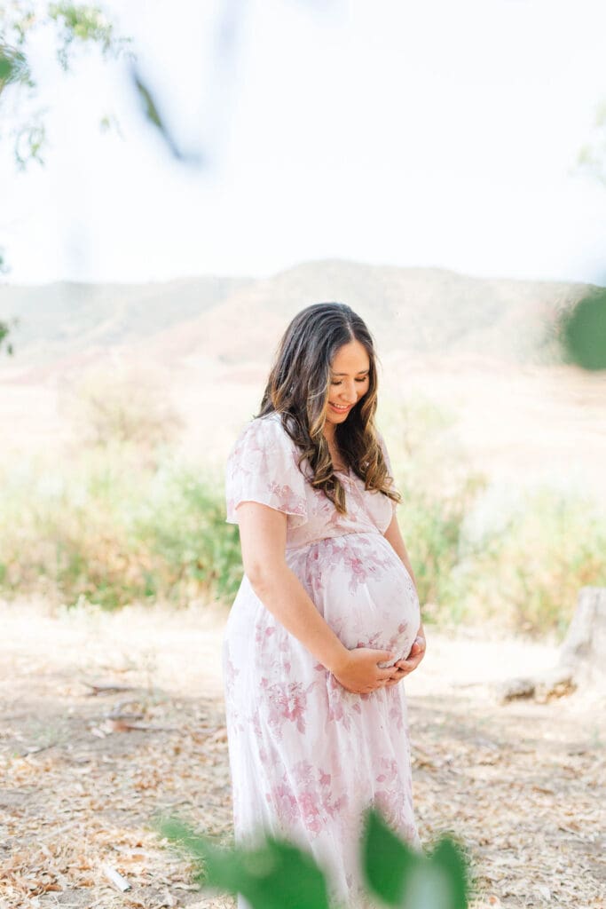 a pregnant woman wearing a floral dress standing in a field and looking down at her belly.