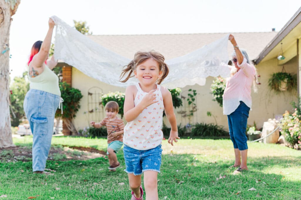 Mom and grandma are holding a blanket up while a little girl runs under it.