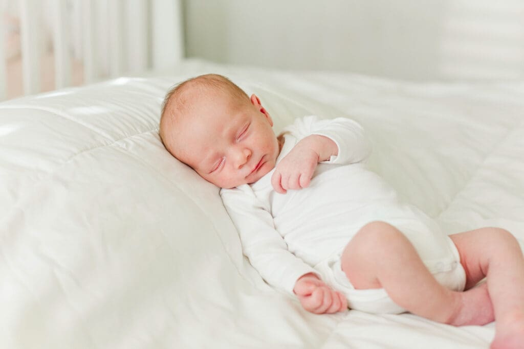 Newborn baby wearing a white onesie laying on a white comforter on the bed with crib in background.