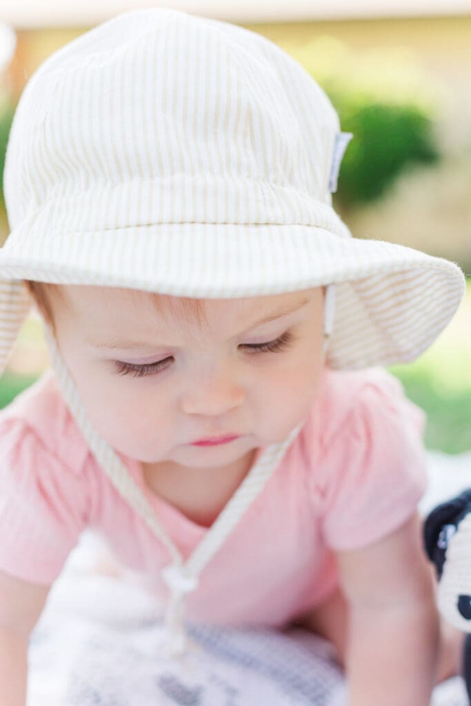 little baby wearing a bucket hat and a pink romper is looking down while crawling.