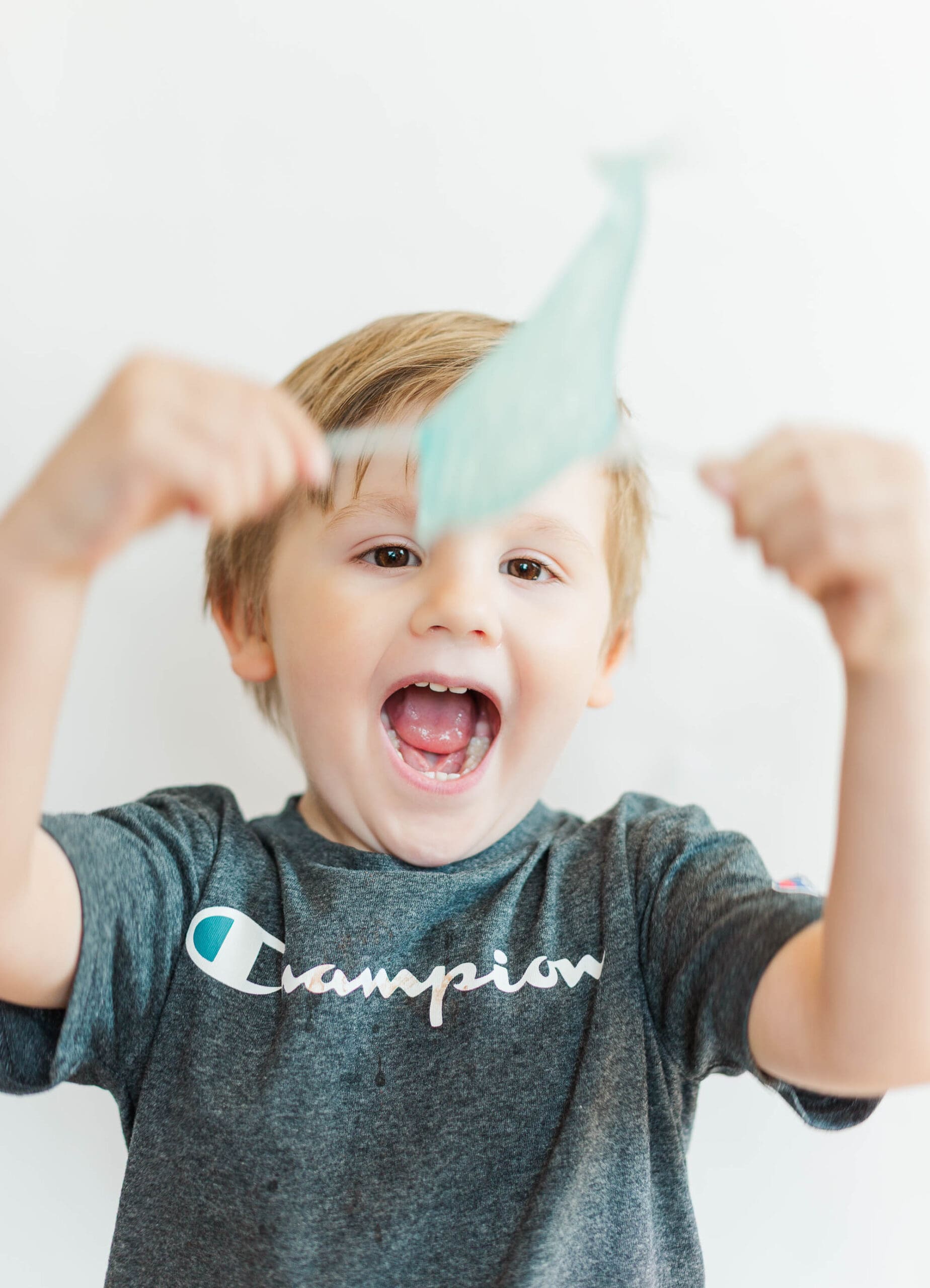 Little boy wearing a grey shirt in front of a white background holding a blue toy and laughing.