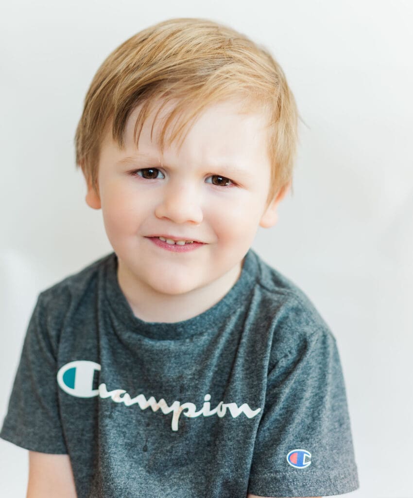 Little boy wearing a grey shirt in front of a white background frowning at the camera.