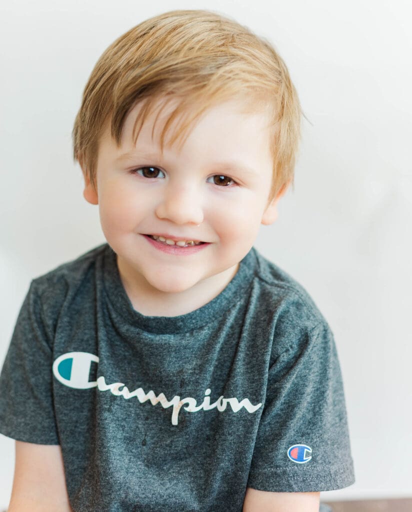 Little boy wearing a grey shirt in front of a white background smiling at the camera.
