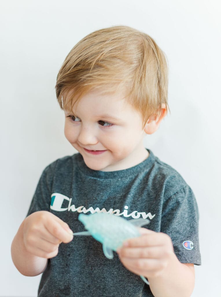 Little boy wearing a grey shirt in front of a white background holding a blue toy, looking off to the side and smiling.