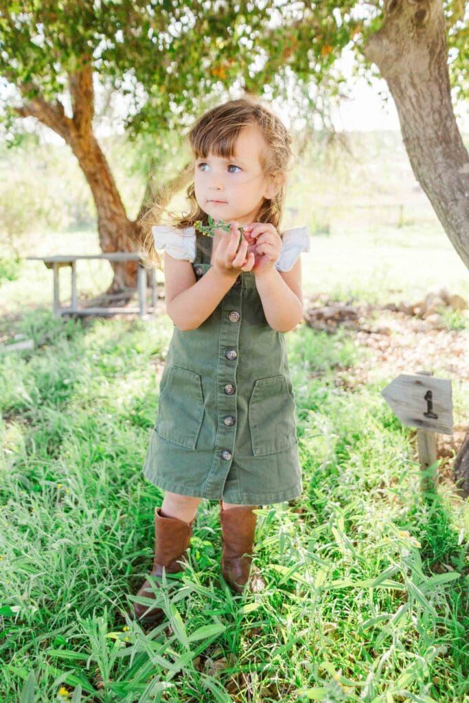 Girl wearing a green overall dress with a white shirt underneath and brown boots. She is standing in a field holding a yellow flower.