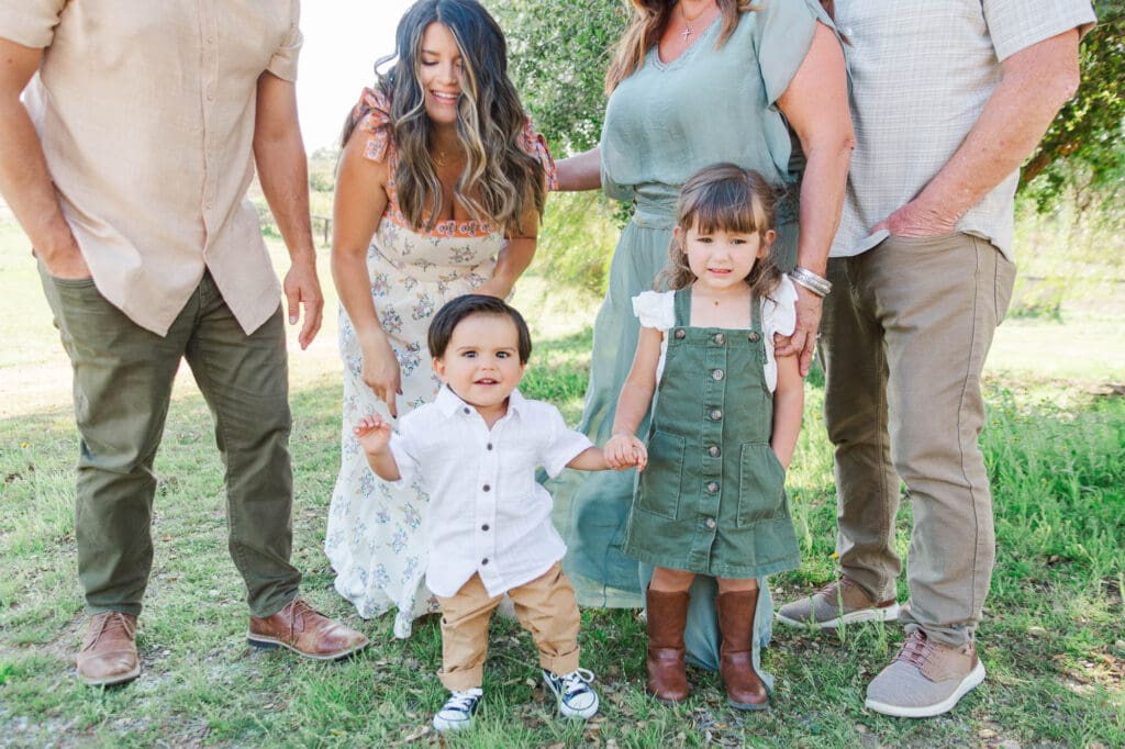 A little boy wearing a white shirt, khaki pants and chucks walking and has a big smile. He is holding his sisters and moms hand and the family is standing in the background.