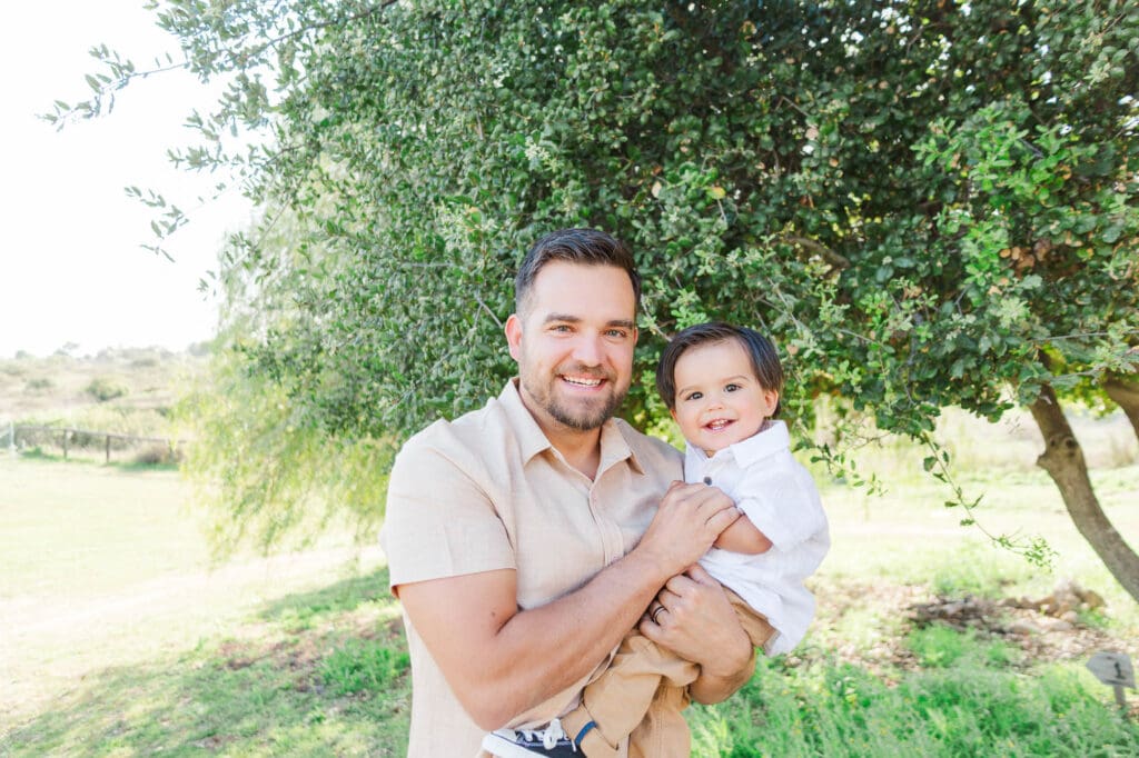 Dad holding his toddler son and they are both smiling at the camera. Dad and son are smiling. Dad is wearing a beige shirt and his son is wearing a white shirt, khaki pants and chucks.